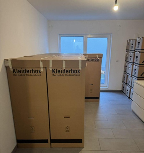 In einem leeren hellen Zimmer mit Fliesenboden stehen mehrere Kleiderboxen aus Karton, dahinter gestapelte Umzugskartons vor einer Fenstertür