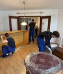 Drei Umzugshelfer arbeiten in einem Wohnzimmer mit Holzboden an einer Schrankwand, im Vordergrund steht ein in Folie gewickelter runder Tisch